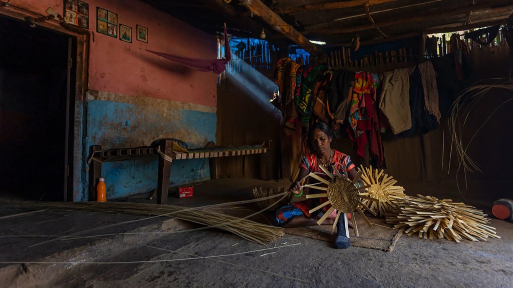A woman sitting on the floor and weaving a bamboo basket. The setting is of a village house.