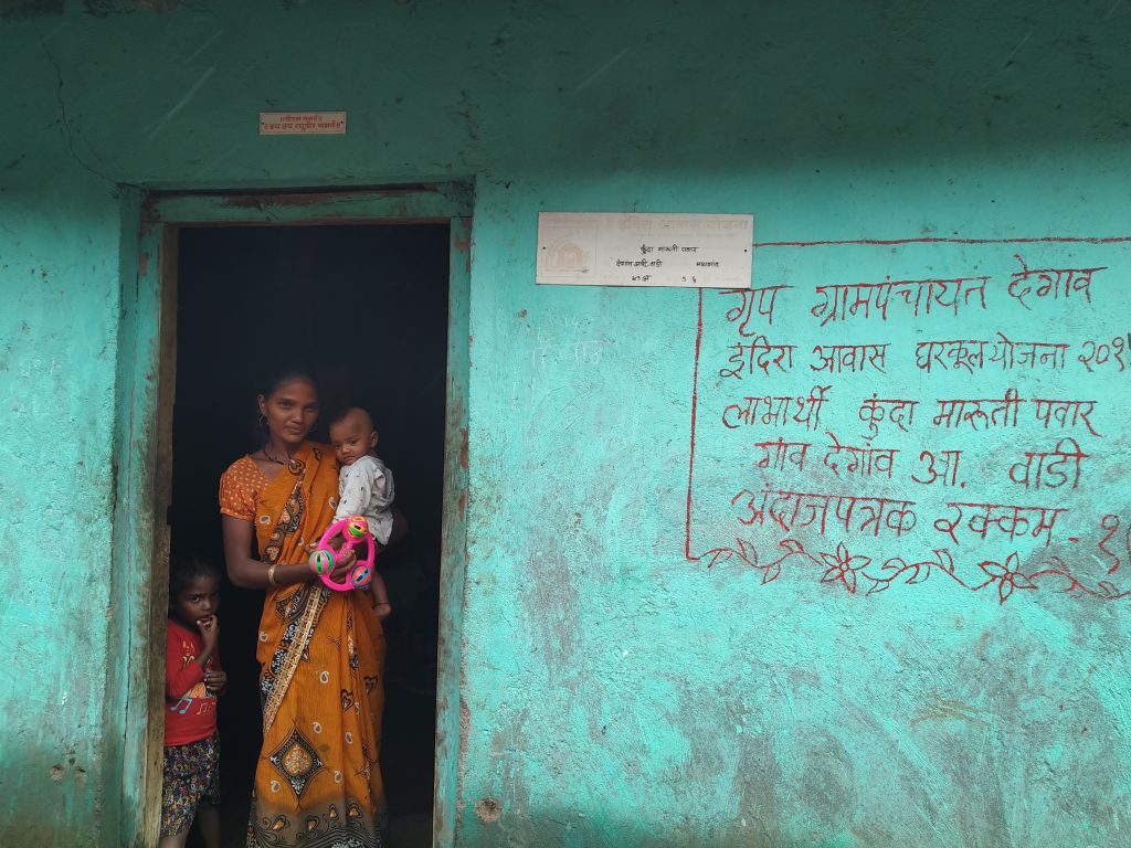 A young woman in an orange saree standing at her house door carrying a toddler. Another toddler is standing next to her. All of them looking at the camera. The front wall of the house is visible - green coloured with the details of the Panchayat name, village name, name of a govt. scheme, name of the woman of the house and amount.
