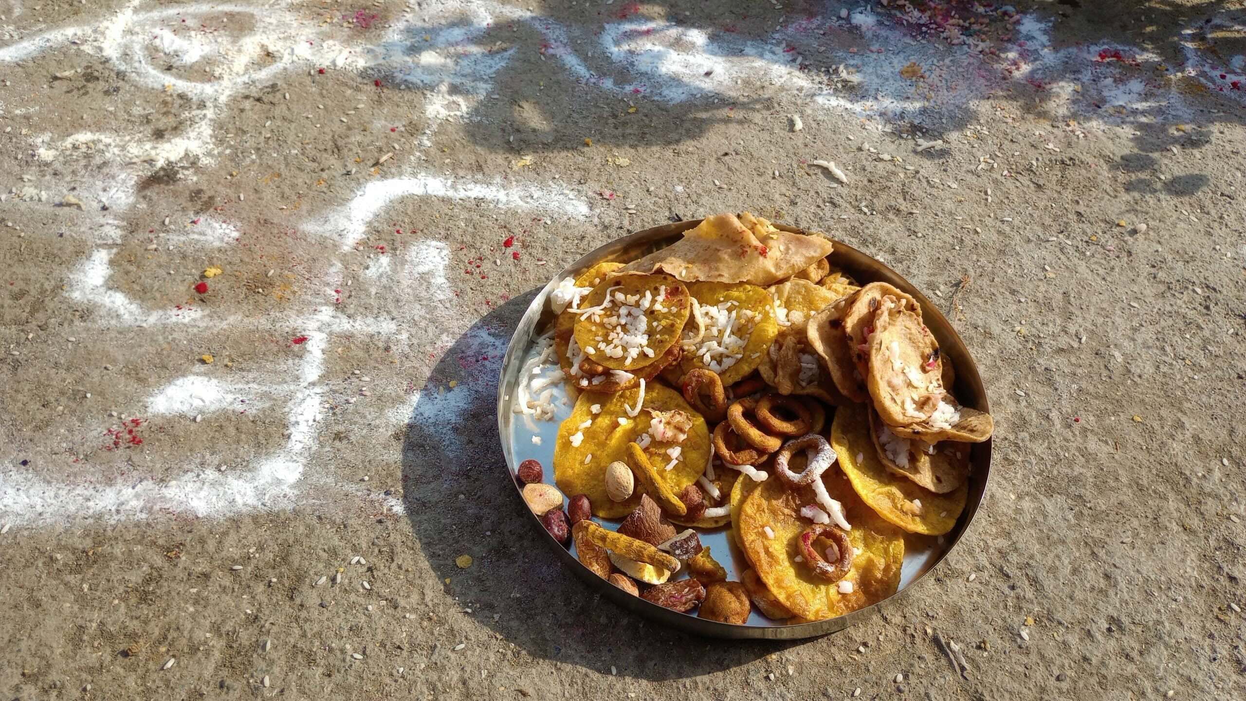 A brass plate filled with various traditional Maharashtrian sweets and snacks, placed on the ground, with colorful rice powder designs in the background.
