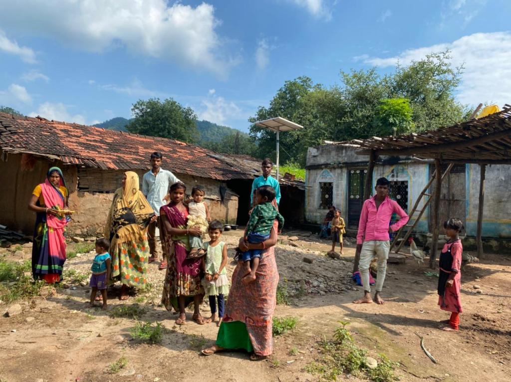 A group of people, including women and children, stands in front of traditional houses in a rural setting in Kotra, Rajasthan, with lush greenery in the background.