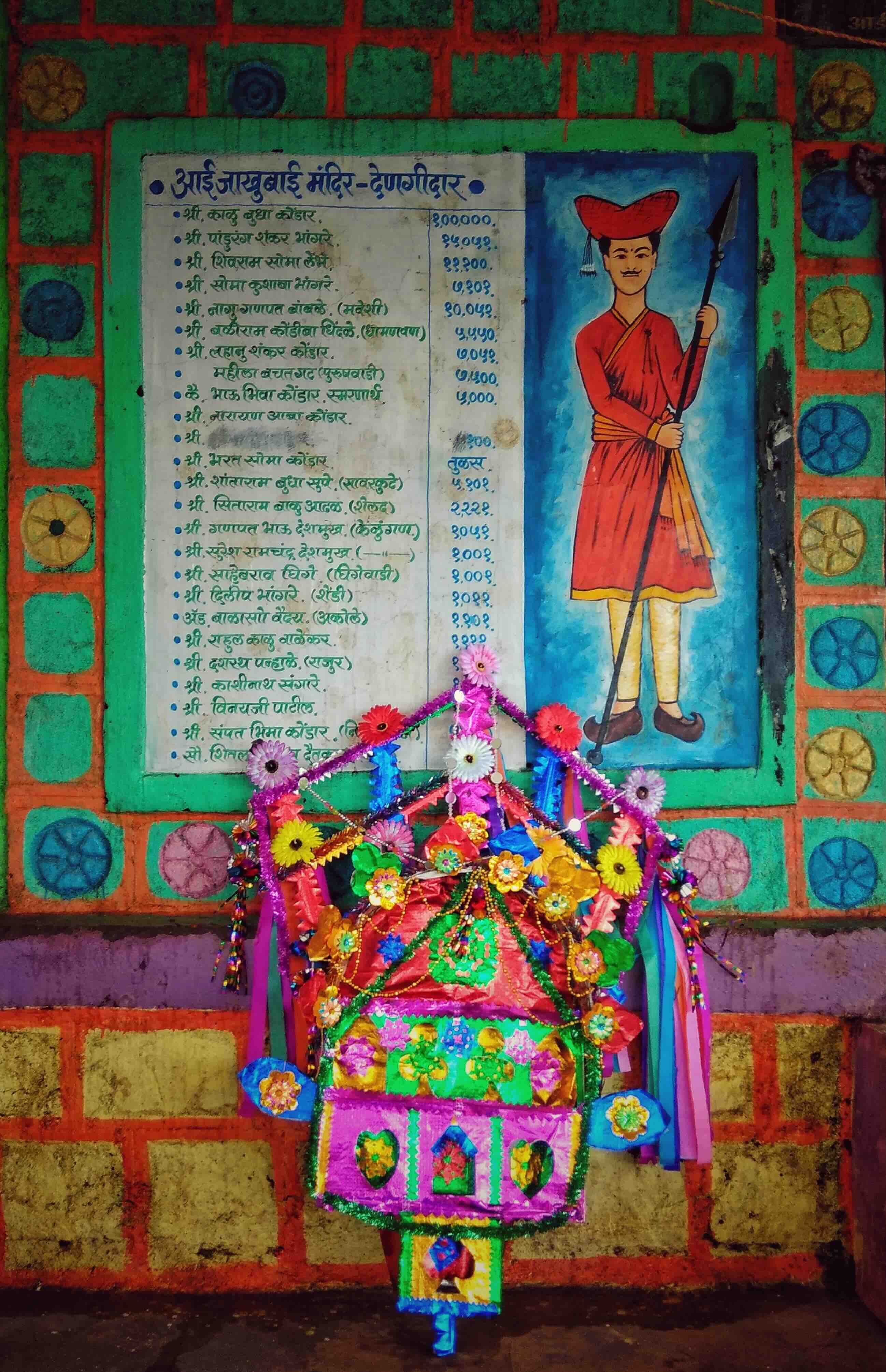 A colorful decorative bullock cart adorned with flowers and ribbons, displayed against a vibrant wall featuring a mural of a man holding a spear and a list of names in Marathi