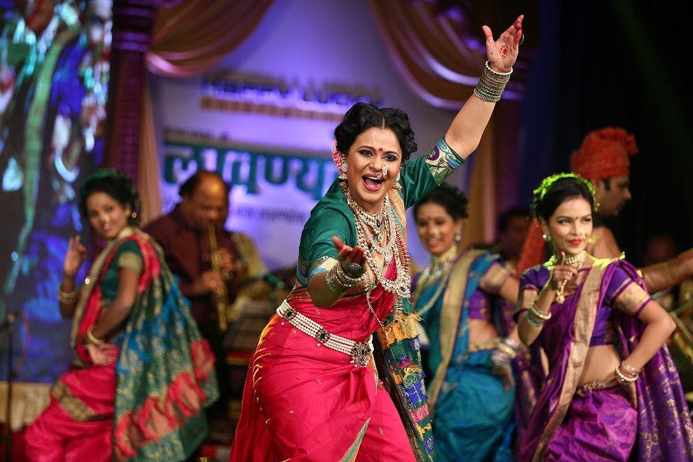 A traditional Tamasha dance performance featuring a female dancer in a colorful saree, joyfully expressing herself on stage, surrounded by other performers in vibrant attire.