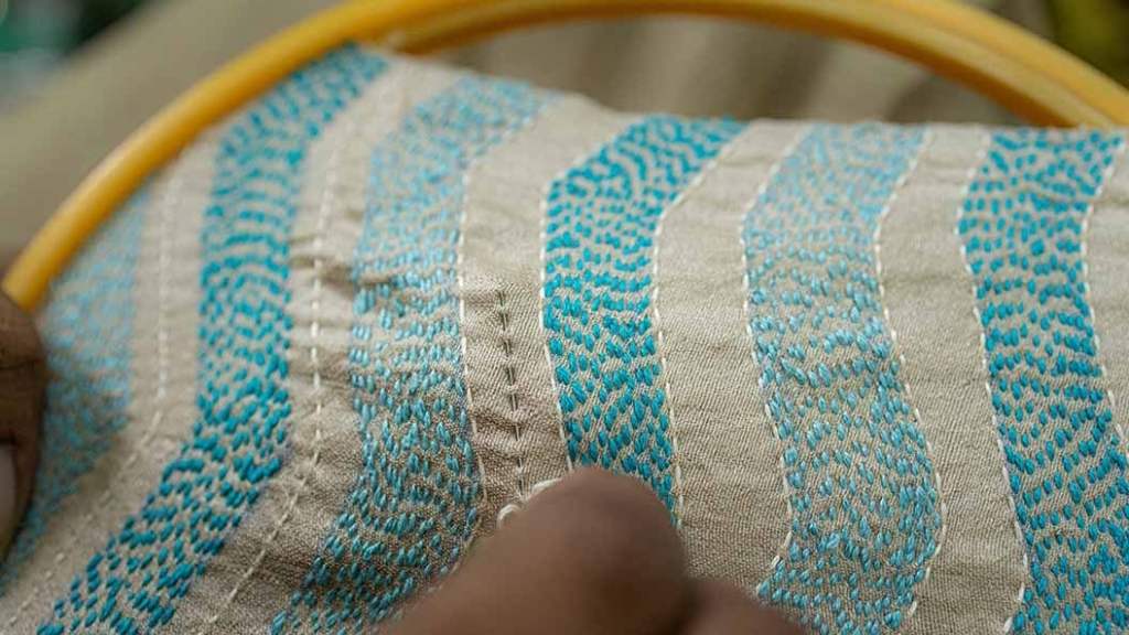 A woman's fingers holding a needle in the foreground. In the background, a cloth held tout between embroidery rings.