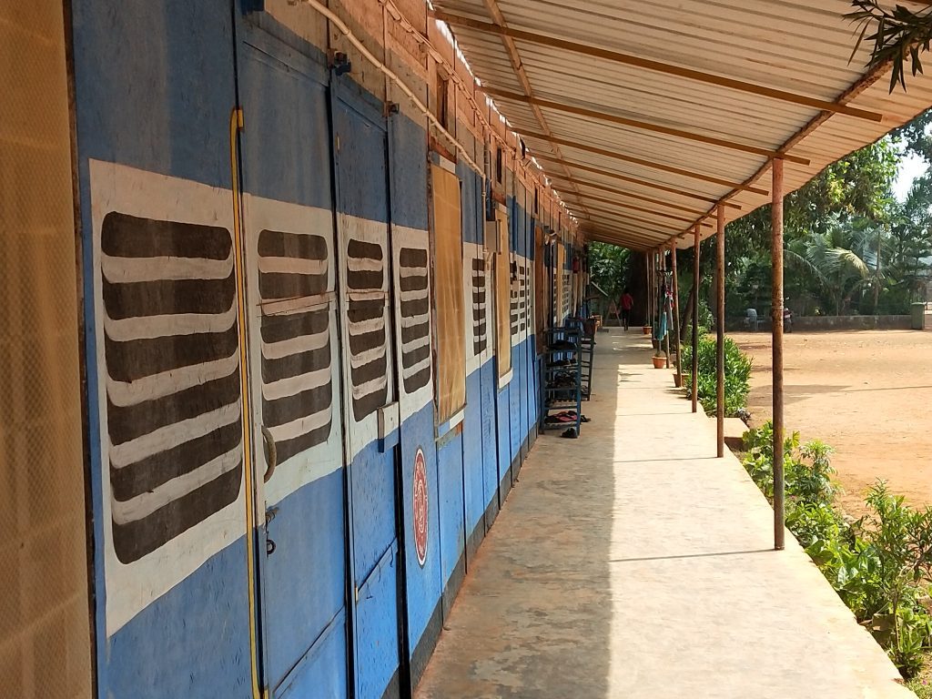 A covered walkway next to a row of blue and white classrooms with open windows and decorative patterns, surrounded by greenery.