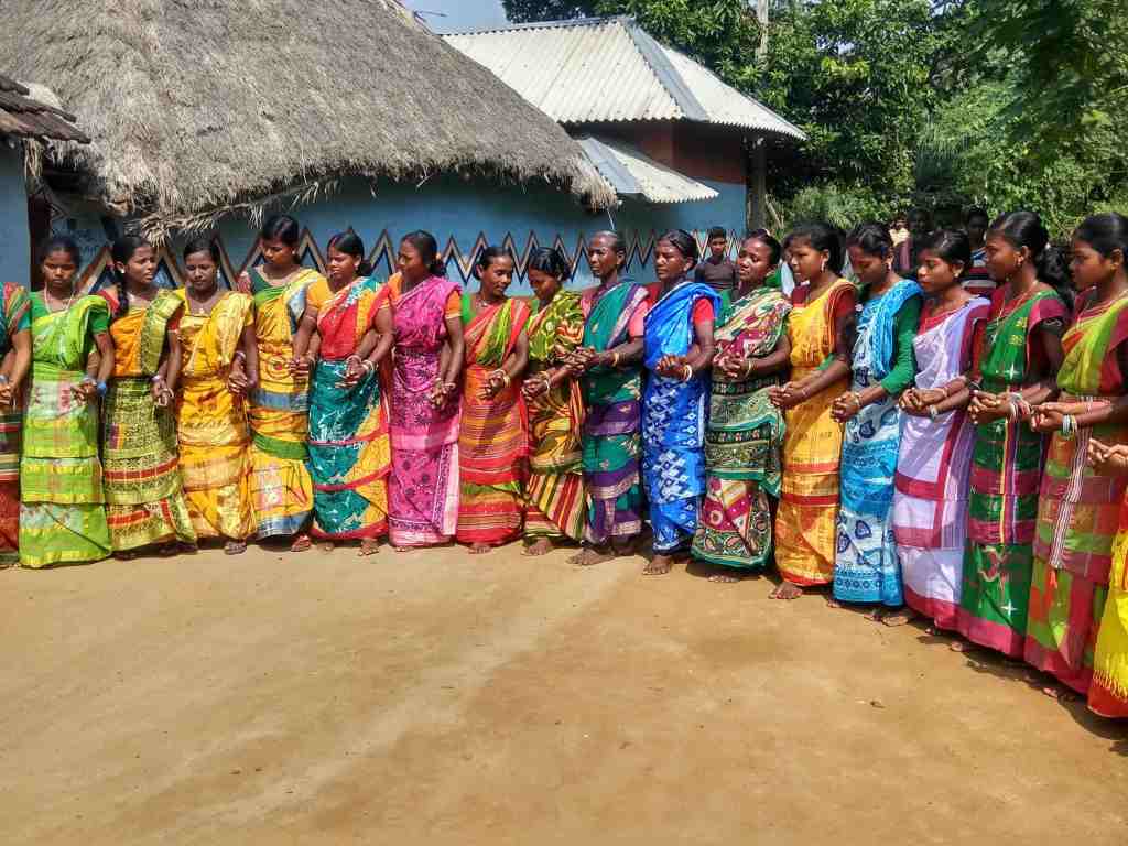 A group of women dressed in colorful traditional sarees standing in a line, holding hands, in a village setting with thatched huts in the background.