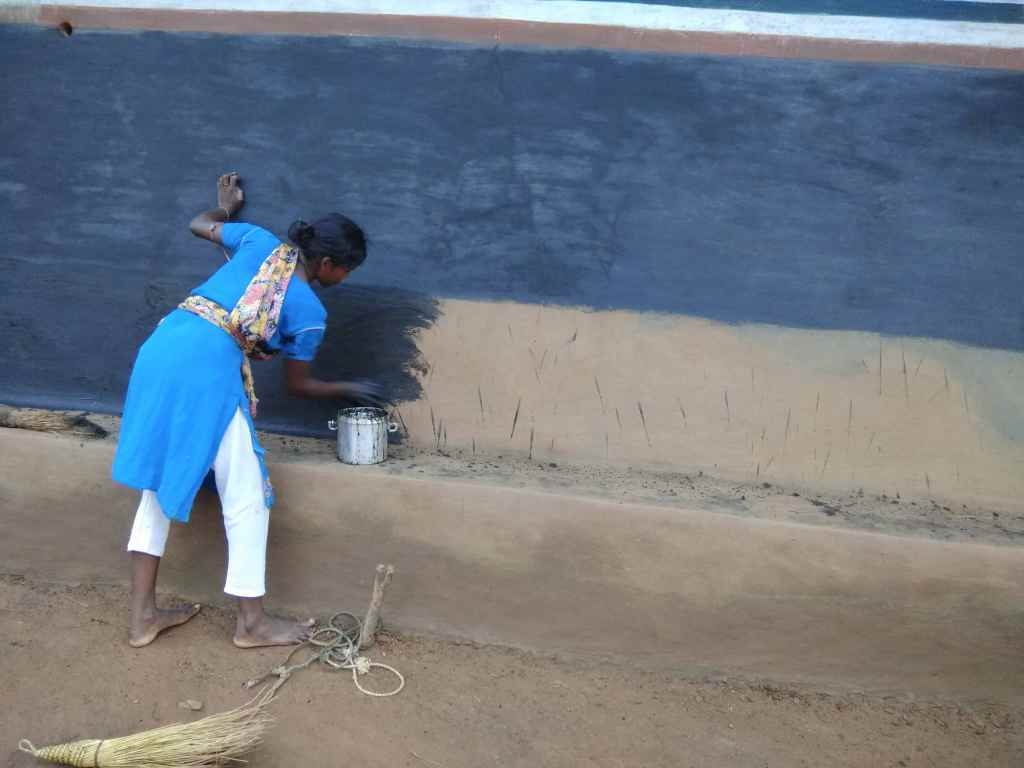 A woman in a blue outfit paints a black surface on a wall while holding a paintbrush, with a container of paint nearby and a broom on the ground.