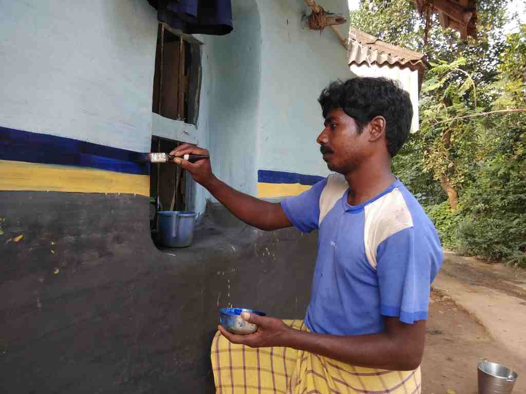 A man painting a wall with blue and yellow stripes while sitting on the ground, holding a paintbrush in one hand and a bowl in the other.