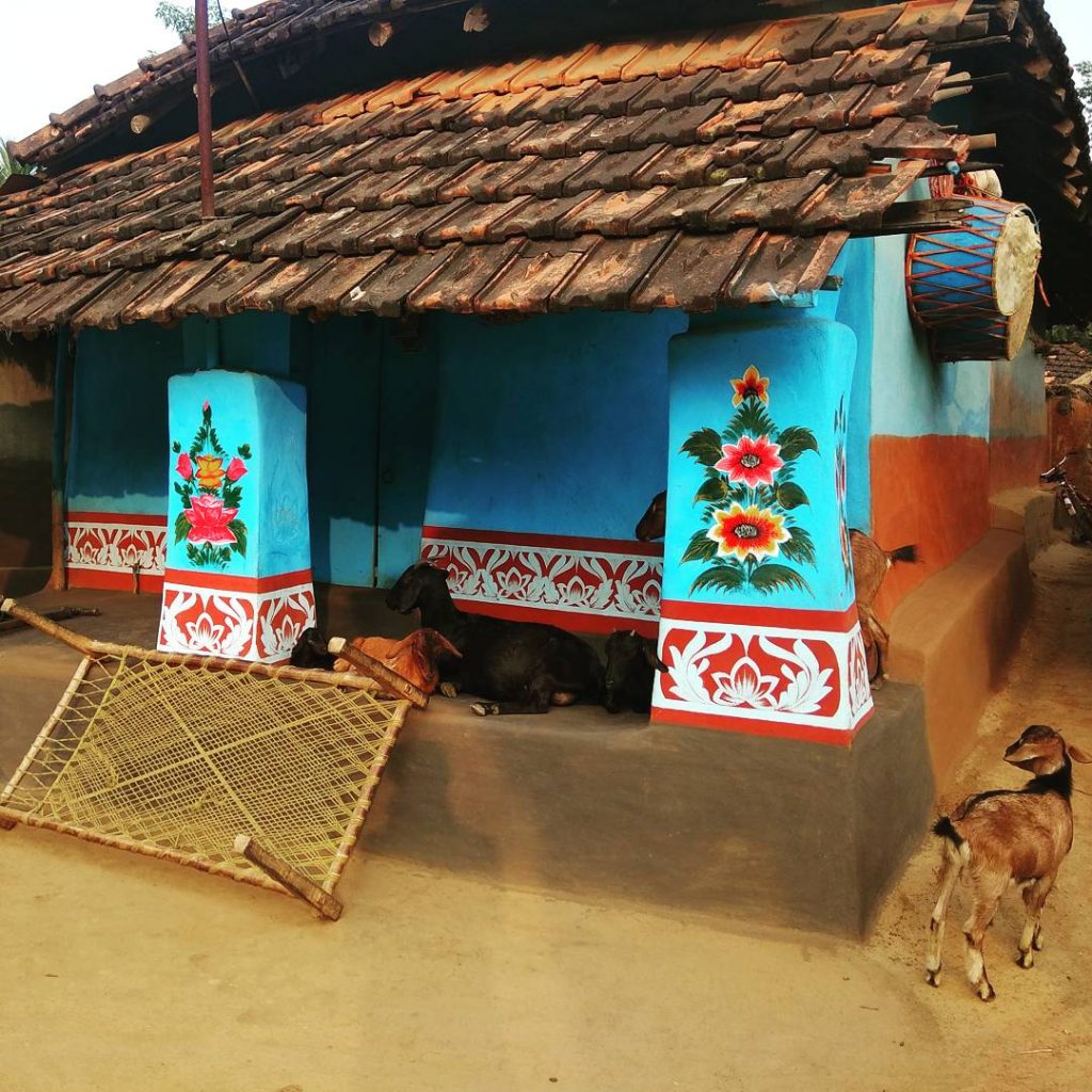 A colorful rural house with blue walls and decorative floral patterns. Bunches of goats are resting around the entrance, and a traditional woven mat is lying on the ground.