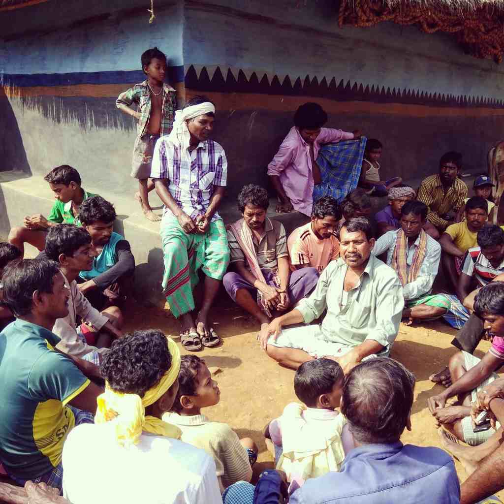 A group of men and children sitting in a circle, engaging in conversation, with some individuals standing nearby in a rural setting.