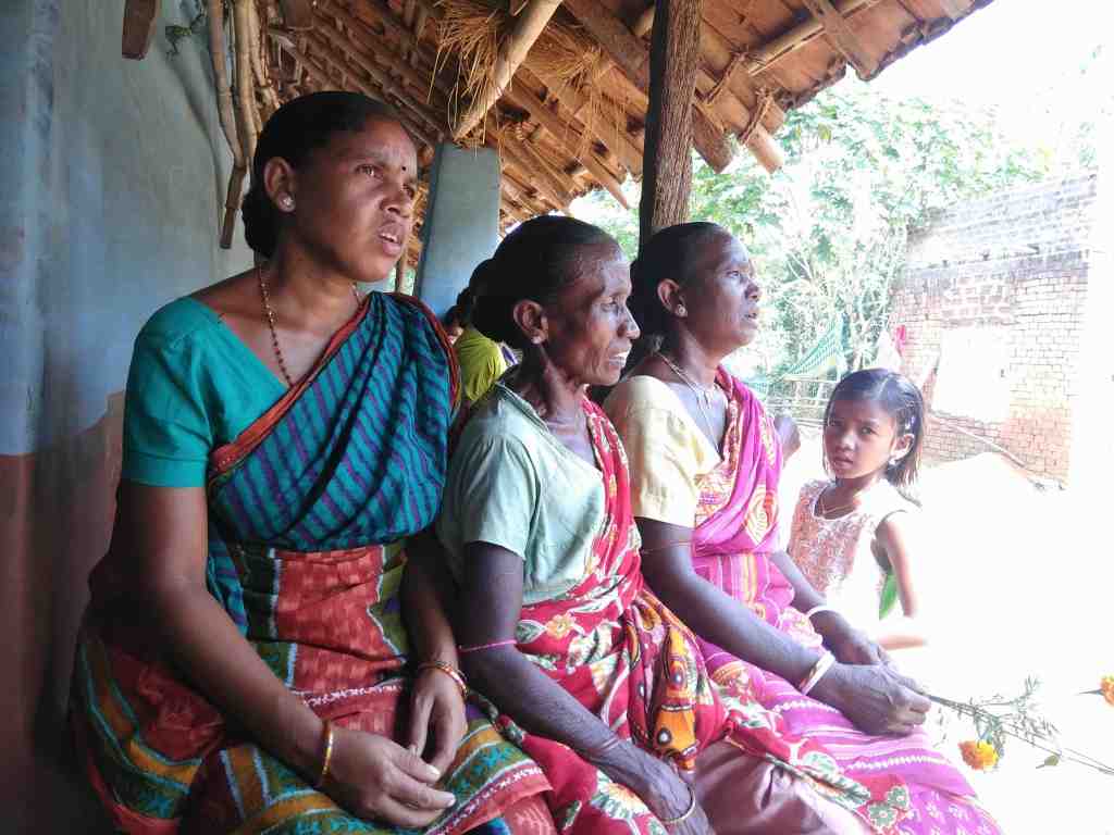 Four women and a girl sitting together in traditional attire, engaged in conversation outdoors near a rustic dwelling.