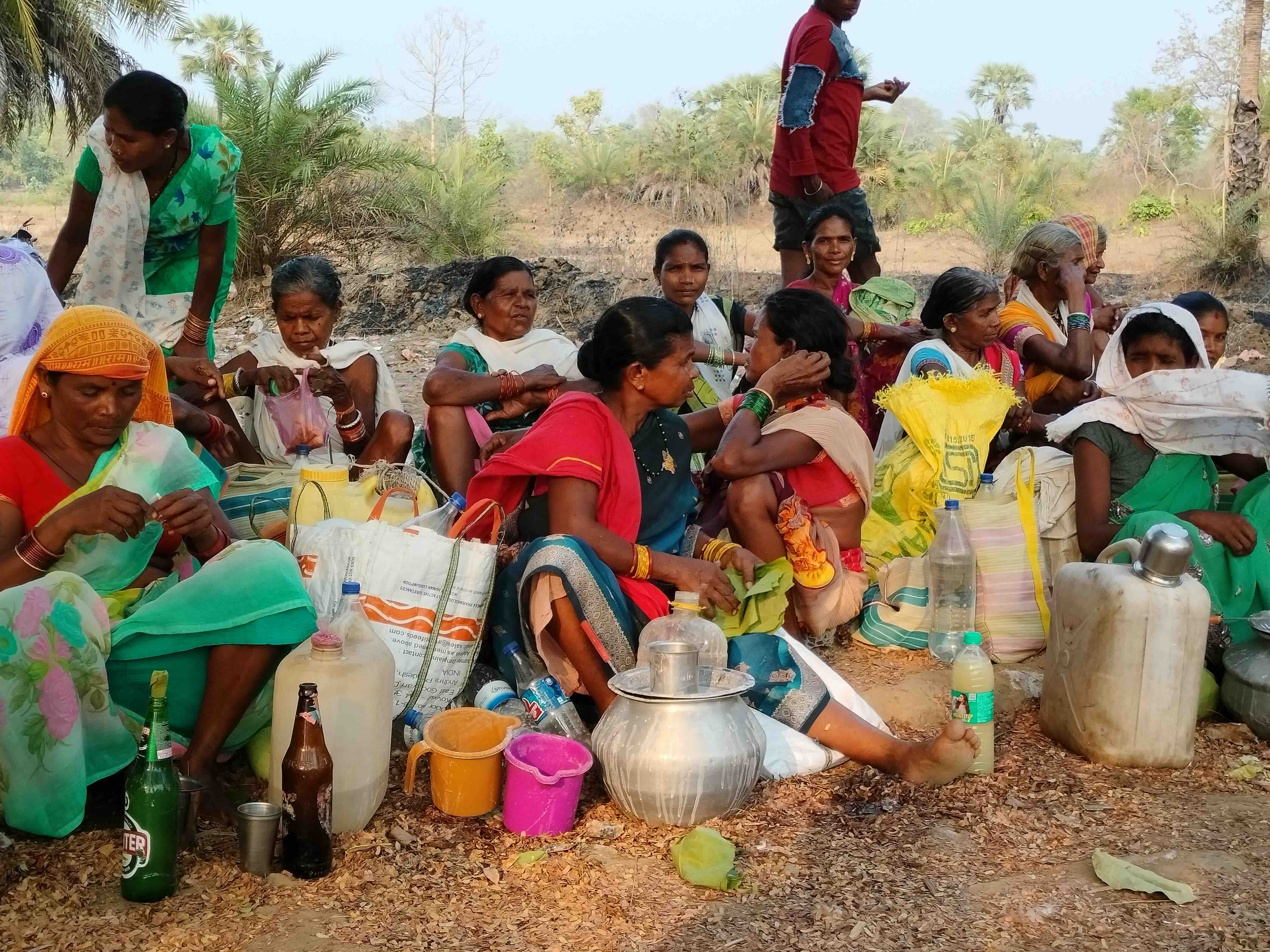 A handful of women, middle aged and old sitting on a ground, with pots, mugs and glass bottles strewn around them.