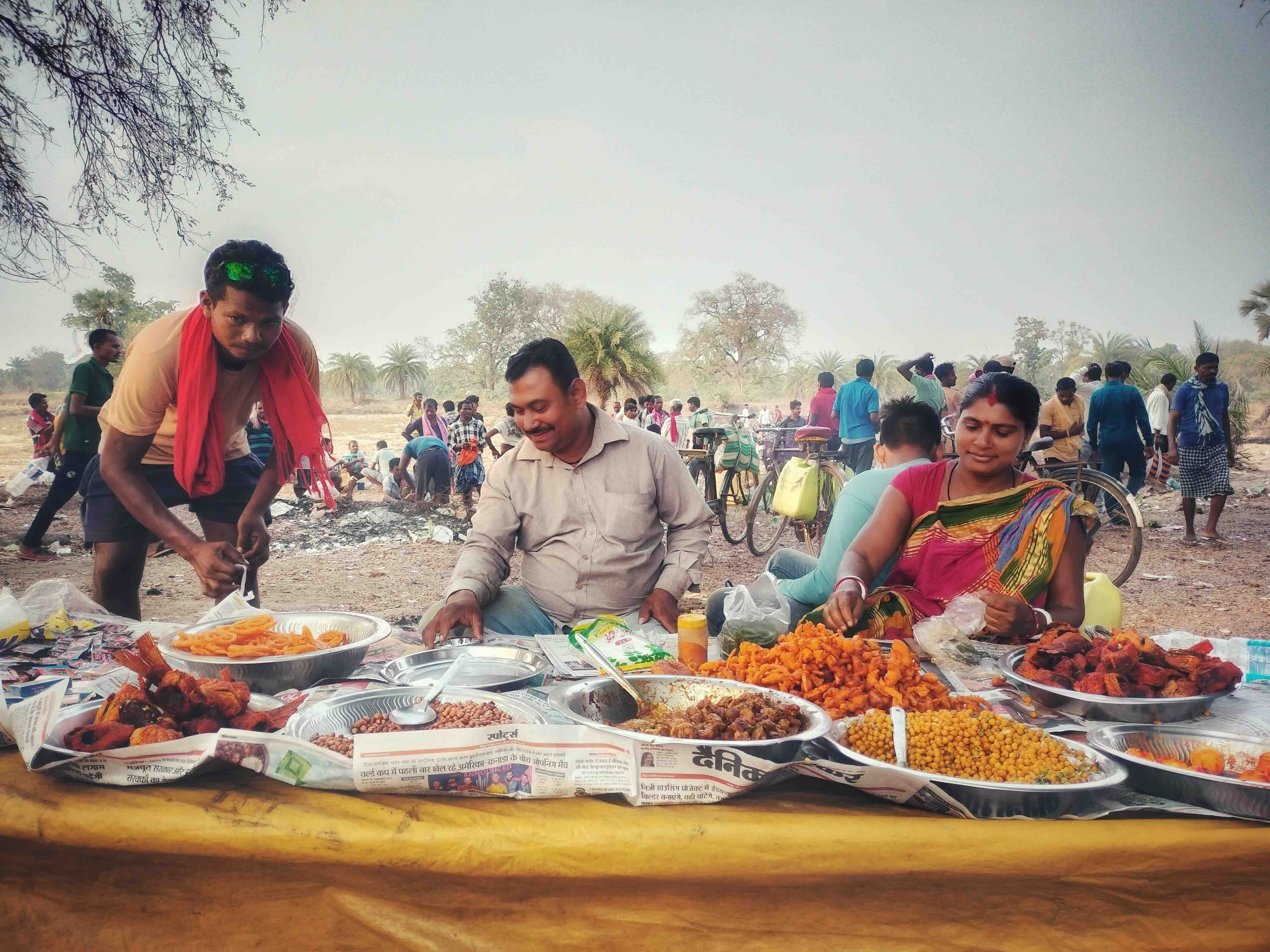 2 men and a woman are sitting, facing the camera, with a string of fried snack items lay ahead of them. Behind them is an open ground, with many men standing in groups.