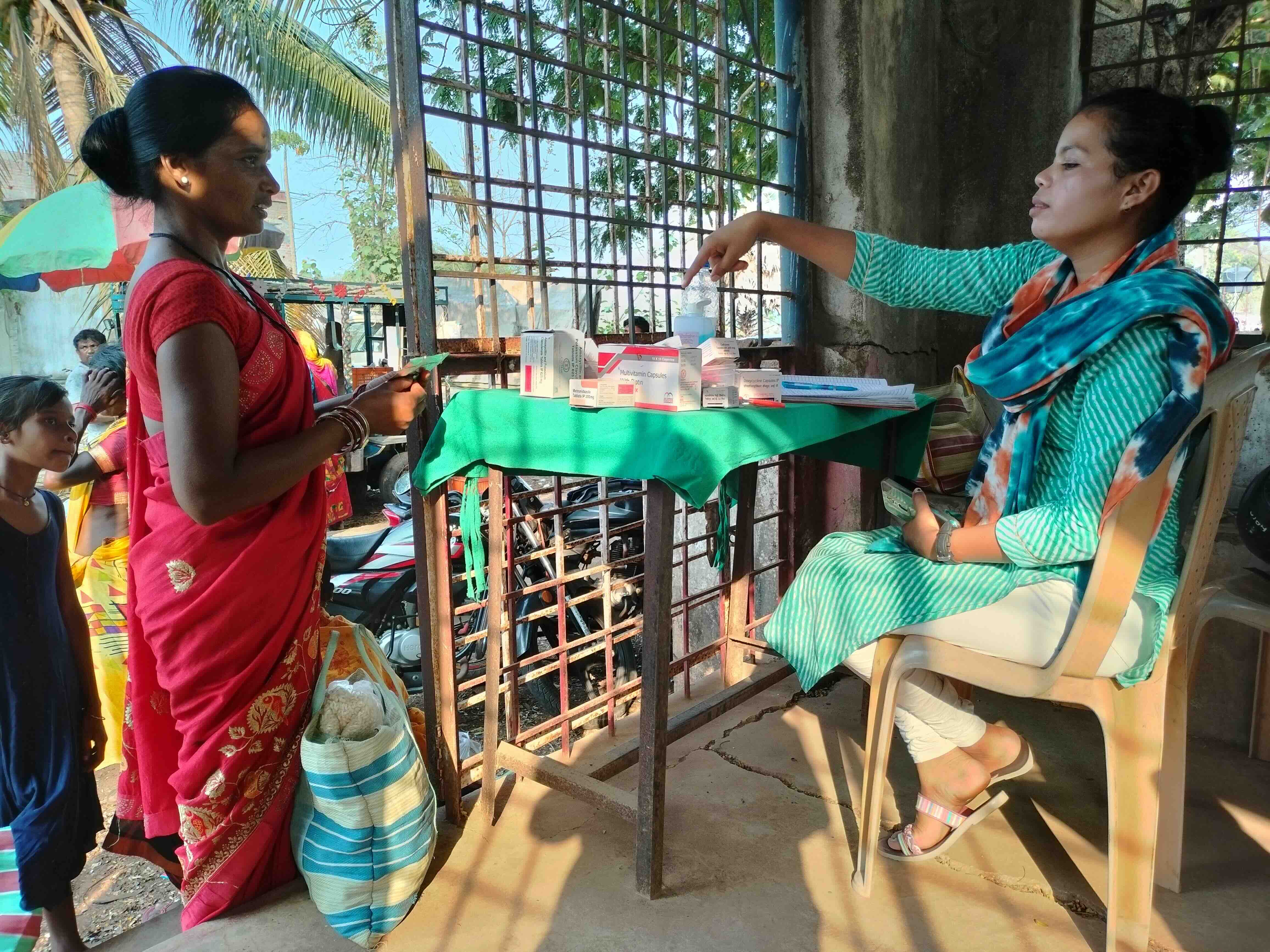 A young woman is sitting on a chair behind a table and handing over a medicine strip to another woman who is standing nearby. The table has some medicine boxes atop.
