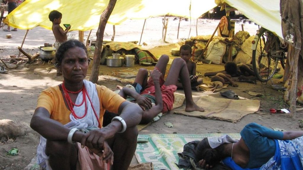 A woman sits on the ground, looking directly at the camera, with tent in the background. Children are visible, some playing and others lying down, while makeshift living conditions are apparent.
