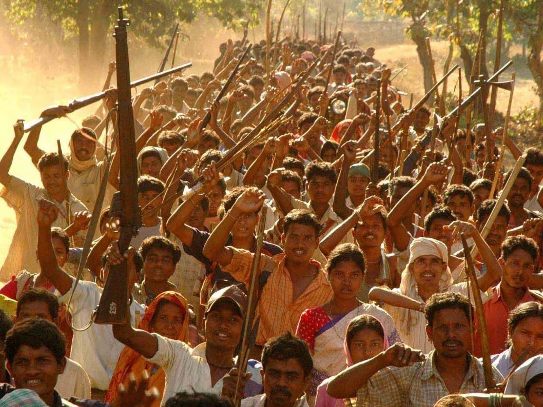 A large group of people marching together, many holding sticks or weapons, with a dusty background and trees lining the path.