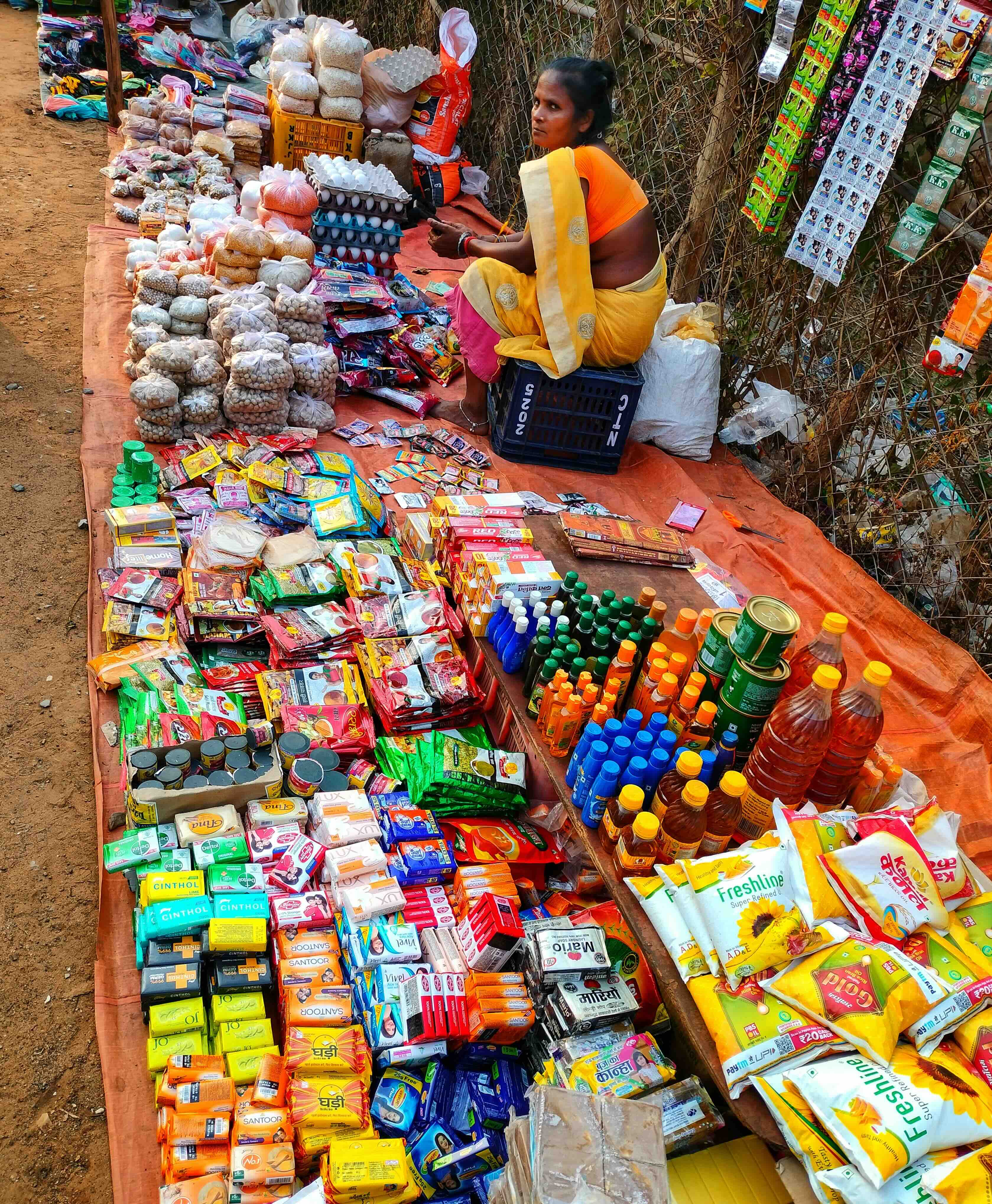 A woman vendor sitting of the ground with a large spread ahead of her. It includes everyday utilities like soap, oil, spices, cleaners, dry snacks, eggs etc.