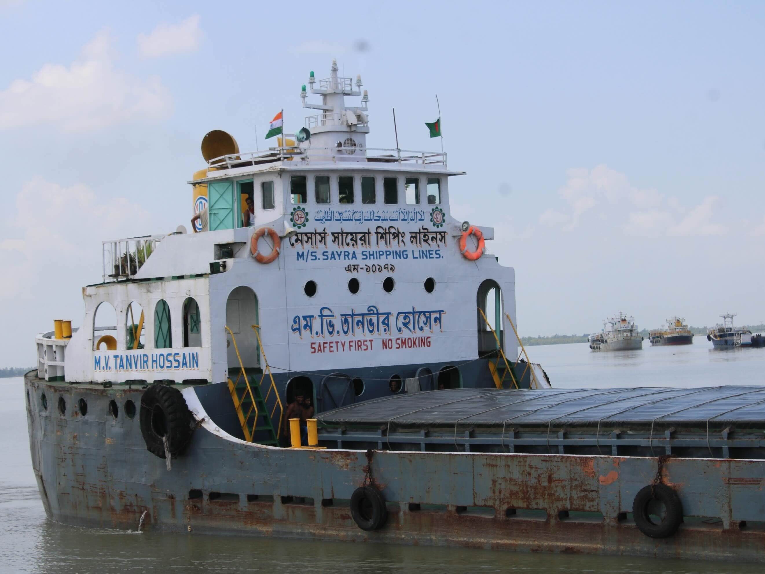 Cargo ship M.V. TANVIR HOSSAIN of M/S. SAYRA SHIPPING LINES on a river. Text on the ship includes: لا إله إلا أنت سبحانك إني كنت من الظالمين, মেসার্স সায়েরা শিপিং লাইনস, M/S. SAYRA SHIPPING LINES., এম-১০১৭৯, এম.ভি.তানভীর হোসেন, SAFETY FIRST NO SMOKING, and M.V. TANVIR HOSSAIN