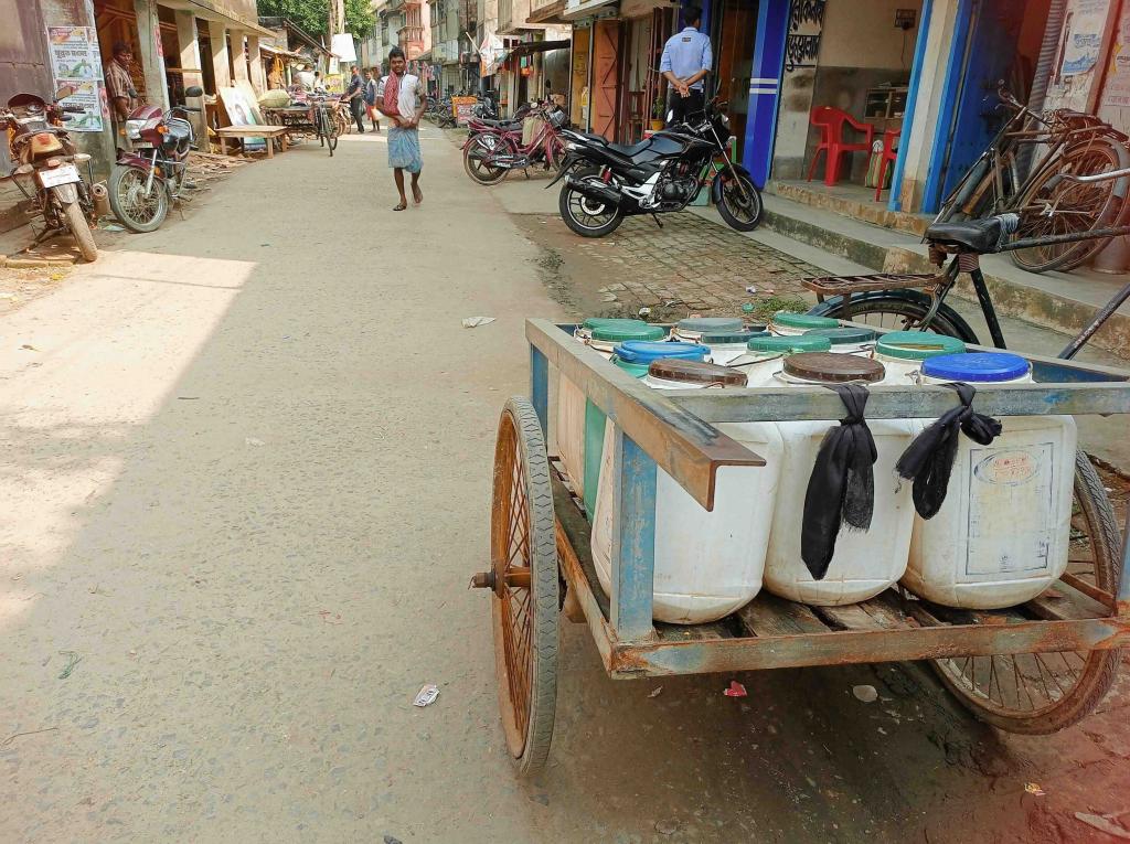 A hand-pulled metal cart filled with large white plastic water containers sits in the foreground of a bustling town street. The containers, featuring an assortment of blue, green, and brown lids, are packed tightly into the cart, which rests on large, thin-spoked wheels. In the background, the sunlit street stretches past parked motorcycles and pedestrians, including a man in traditional attire walking toward the camera and another standing near a blue-fronted shop. The image captures a candid slice of daily life, with a warm, dusty color palette that emphasizes tje practical nature of local goods transport.
