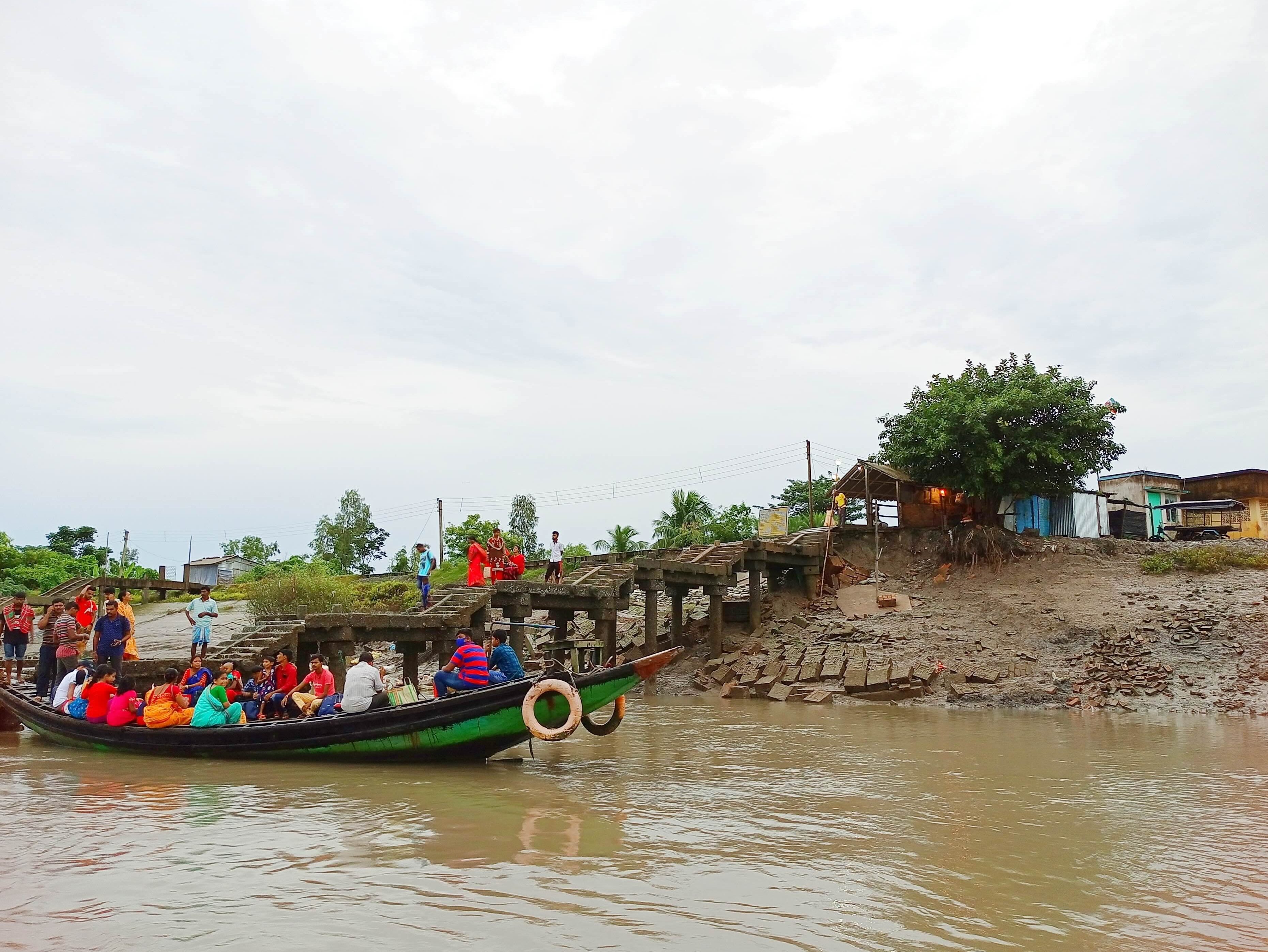People boarding a crowded boat at a concrete landing on a muddy riverbank