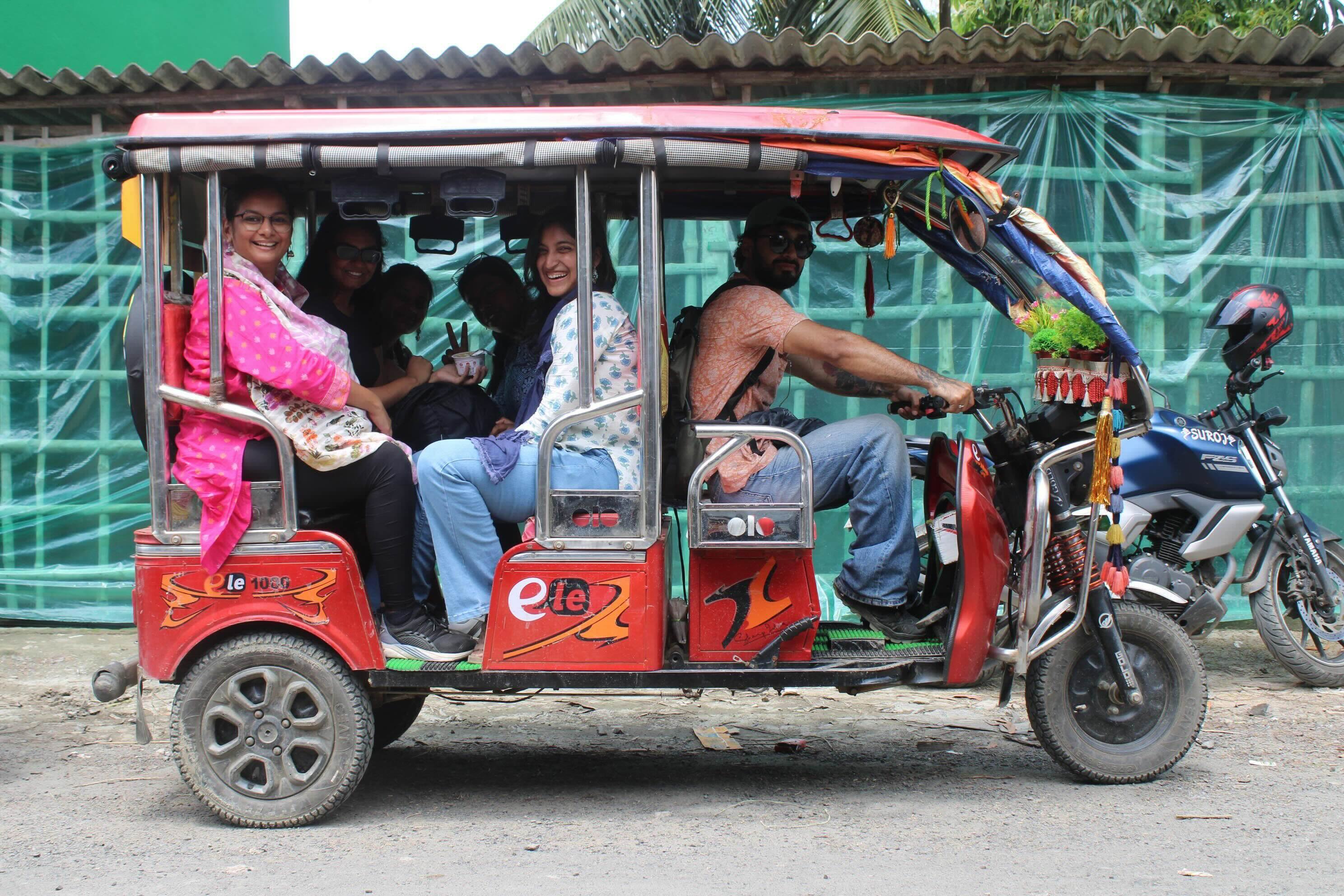 A group of smiling young people is captured in a candid moment while riding in a vibrant red electric rickshaw, often called a toto, on an unpaved road. The rickshaw, decorated with colorful tassels and metallic framing, carries four women in the back and a man wearing sunglasses at the helm, all looking toward the camera with joyful expressions. The scene is set outdoors against a background of green mesh fencing and a parked blue motorcycle. The image's composition features a full side-view of the vehicle, emphasizing the bright red color of the rickshaw and the diverse, colorful attire of its passengers.