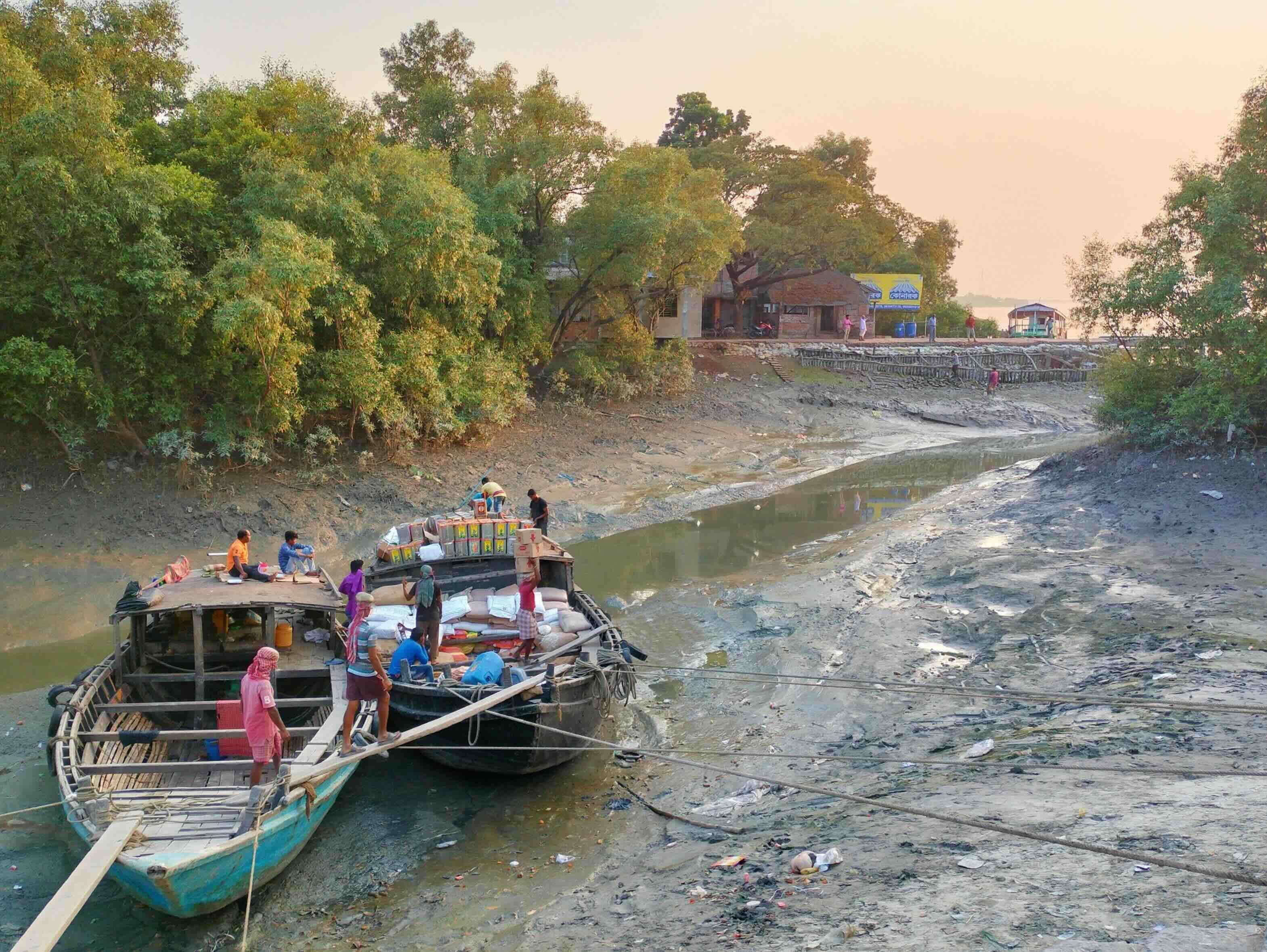 Workers unloading goods from wooden boats onto a muddy riverbank at low tide.