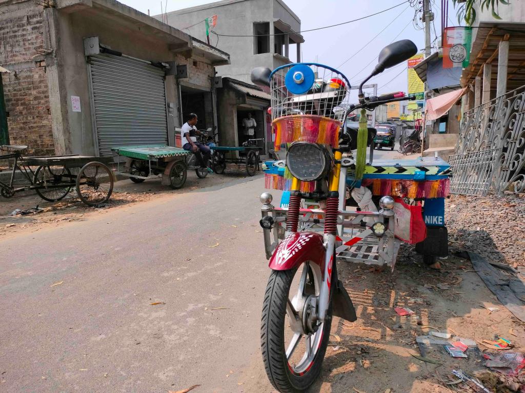 A colorful, customized electric tricycle with a flatbed is parked on a paved road. The vehicle is heavily decorated with vibrant multi-colored tassels, a blue circular horn on its wire front basket, and a red front fender featuring white graphic patterns. In the background, single storey buildings with metal shutters and a person sitting on another transport cart create a sense of daily life, while a Trinamool Congress political flag flies from a nearby roof. The bright, natural daylight highlights the vivid colors of the tricycle's decorations against the neutral tones of the dusty road and surrounding architecture.