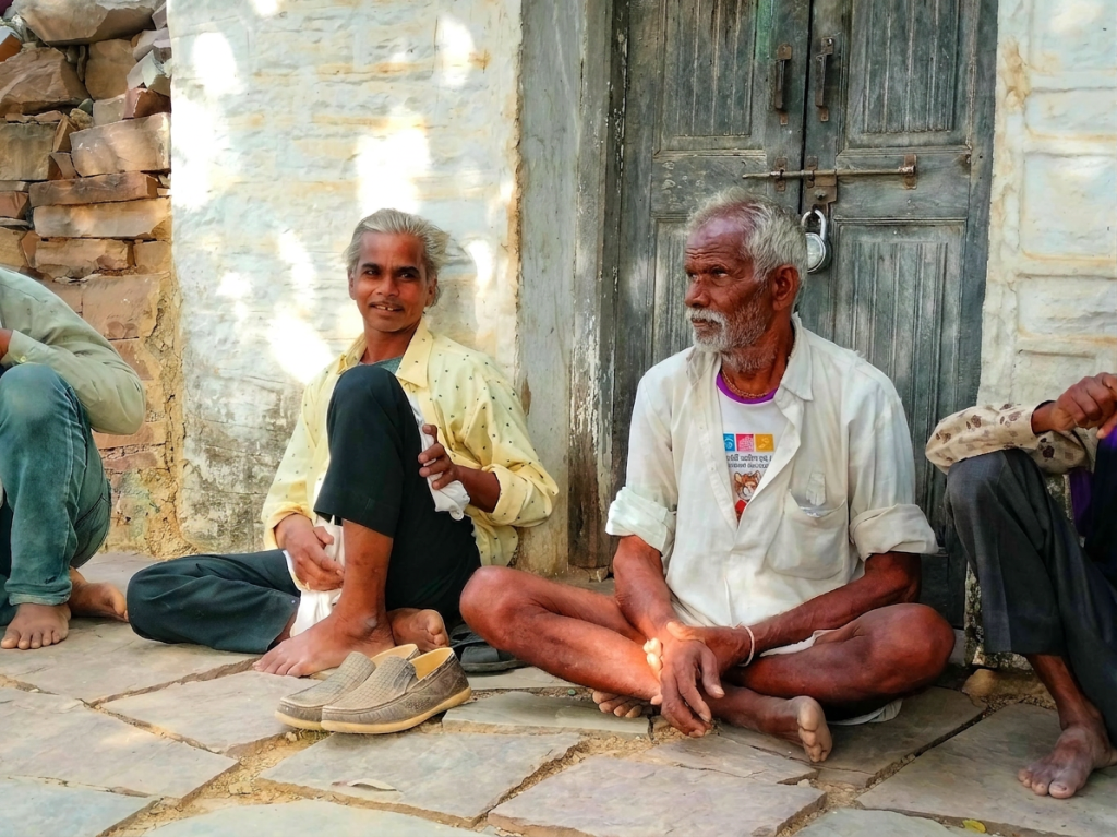 Two men sitting barefoot on a stone floor outside a locked door, one smiling and the other looking to the side