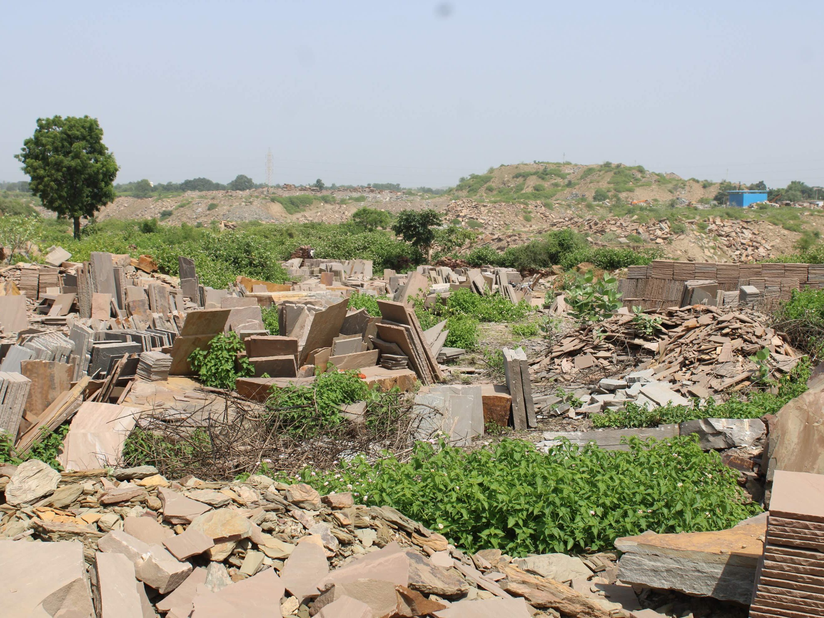 Stacks of stone slabs and tiles in an outdoor storage area with scattered greenery and hills in the background